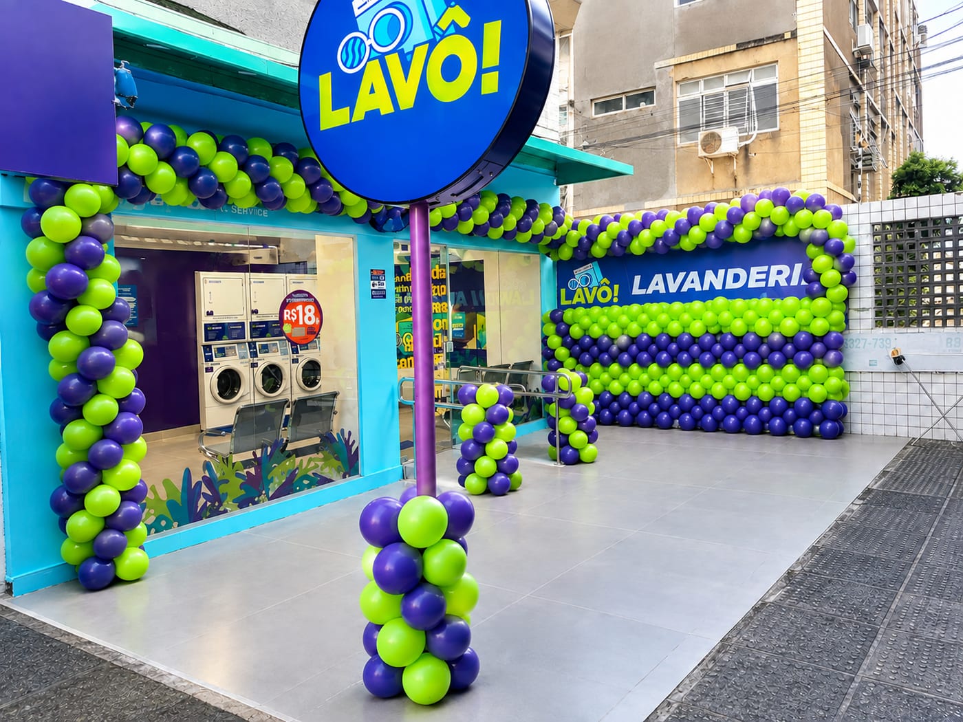 Fachada da Lavô Pina, uma loja azul com o letreiro redondo LAVÔ!, a vitrine com as máquinas e a entrada enfeitada com balões roxos e verdes