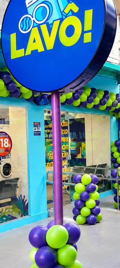 Letreiro redondo azul com a marca LAVÔ! no poste roxo da calçada, enfeitado com balões
